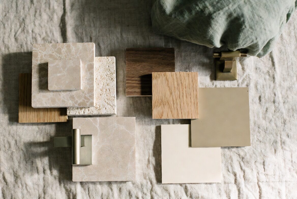 An interior designer's material palette — stone and tile samples, wood veneers, paint swatches in bone and putty, brass hardware, and textiles.